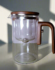 Clear glass tea pot with wooden handle and lid on a light green surface.