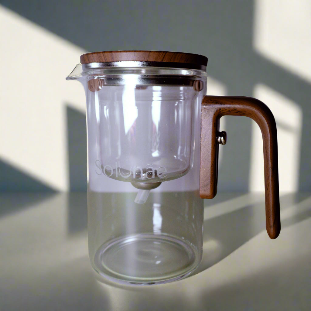 Clear glass tea pot with wooden handle and lid on a light green surface.